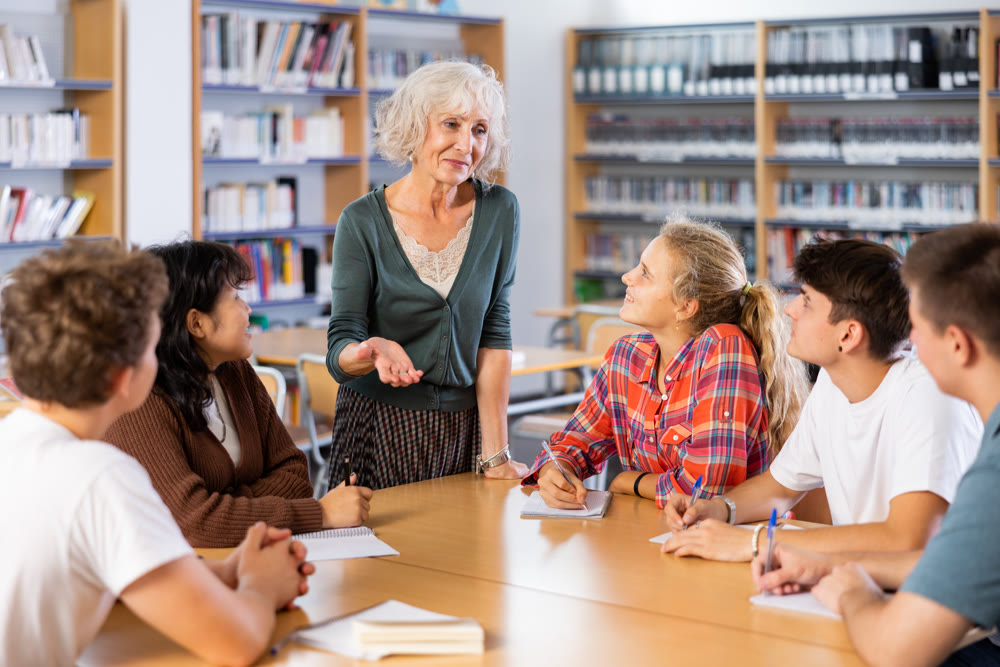 Mature female teacher working with friendly group of schoolgirls and schoolboys studying in library