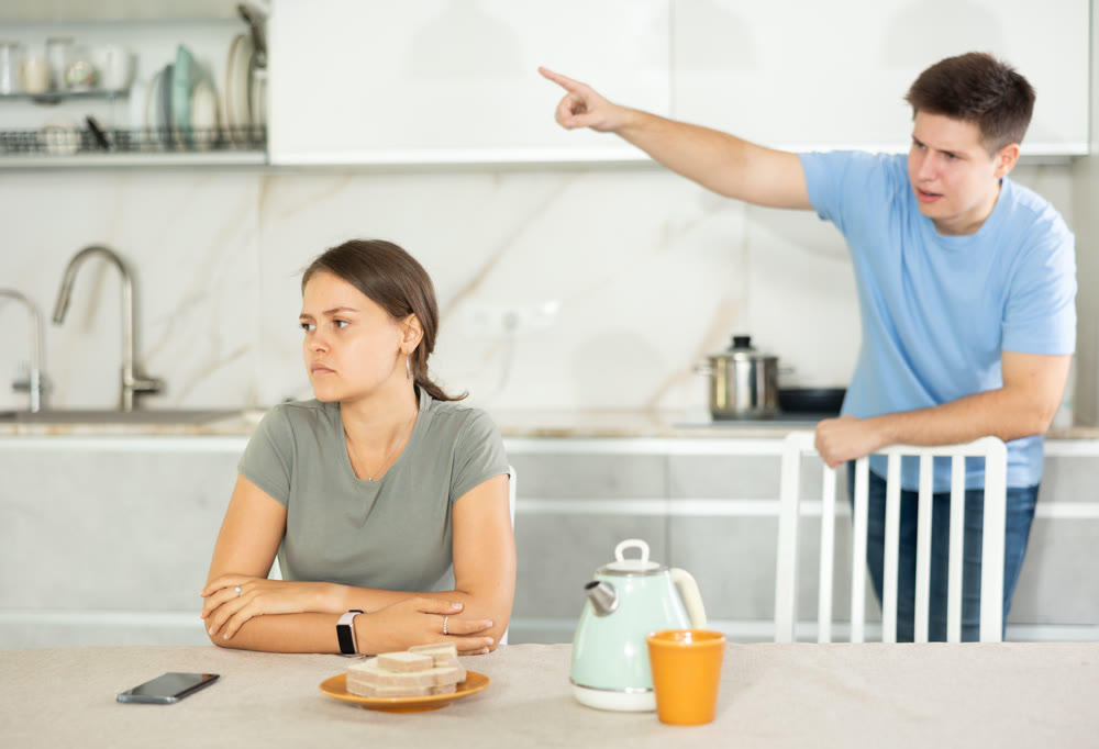 Young girl sitting at the table while her boyfriend is quarreling with her standing behind