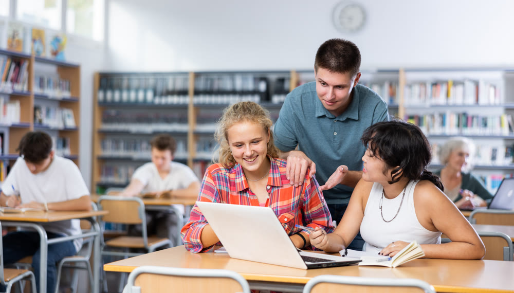 Smiling boy helping positive pupils research, working with laptop in library