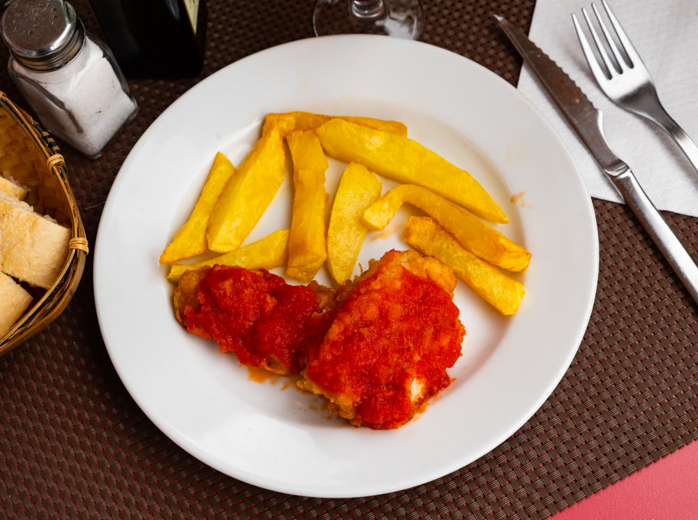 There is large slice of fried cod on plate, covered with tomato sauce and French fries.