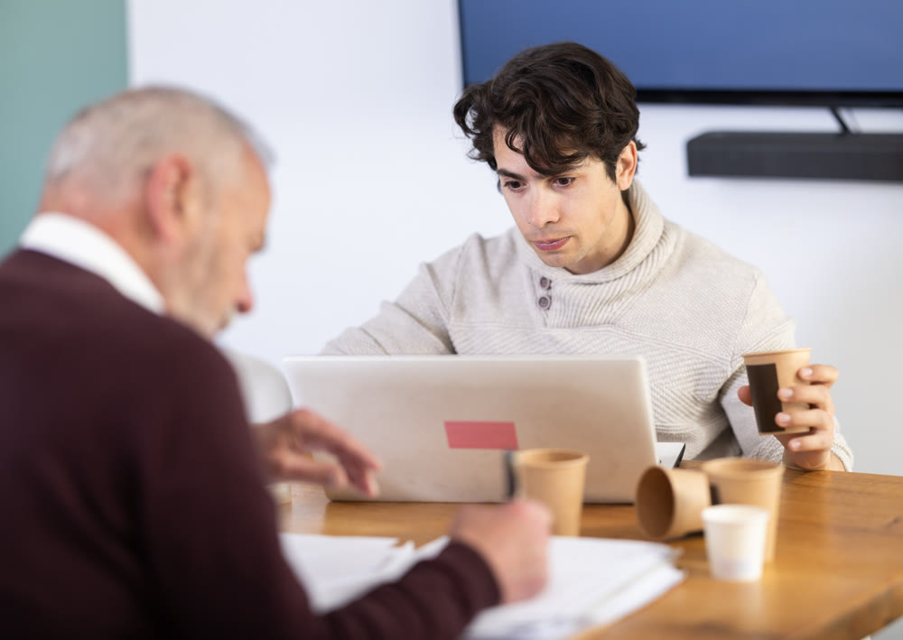 Young man working on laptop, elderly man takes notes on documents
