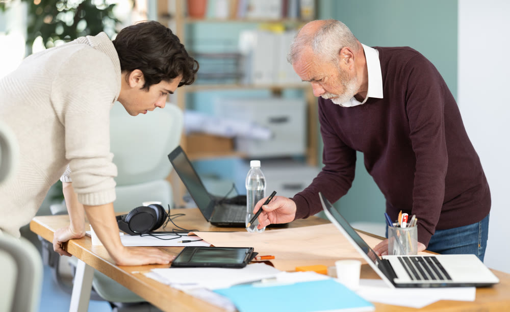 Two men are bent over drawing, reviewing and discussing development of construction.
