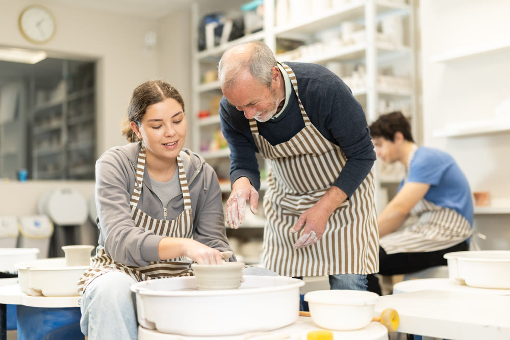 Eldery man helping young woman shape pottery on wheel