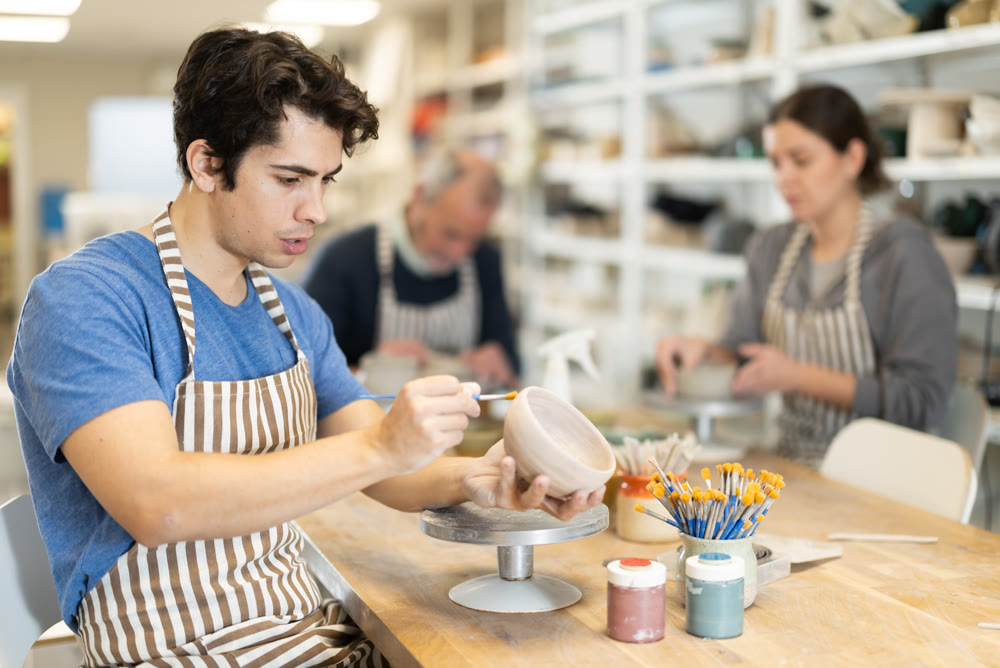 Young man applying coating with brush on ceramic piece