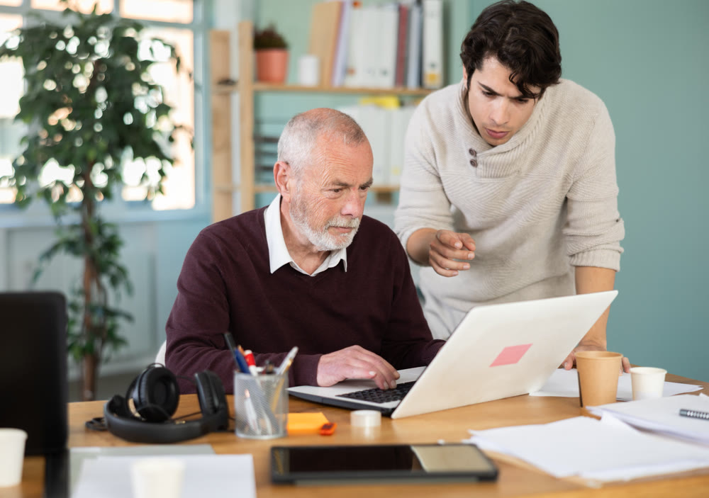 Young man helping elderly male colleague work on laptop
