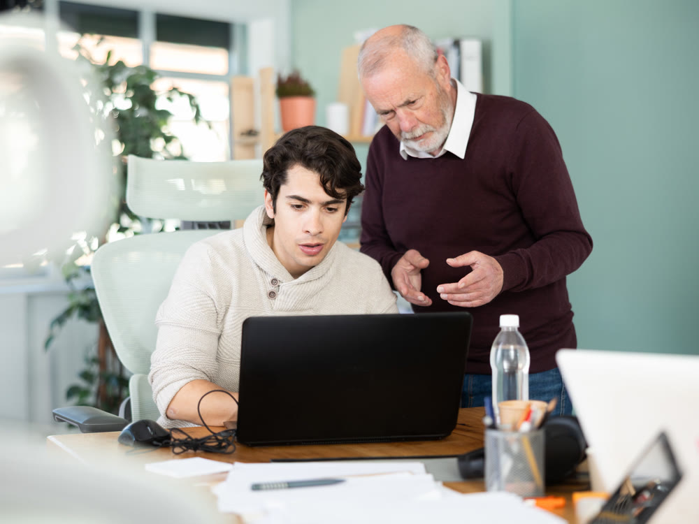 Elderly man helping young male colleague work on laptop