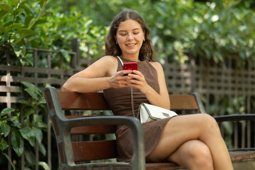 young woman sits on a bench in summer with a phone in her hands