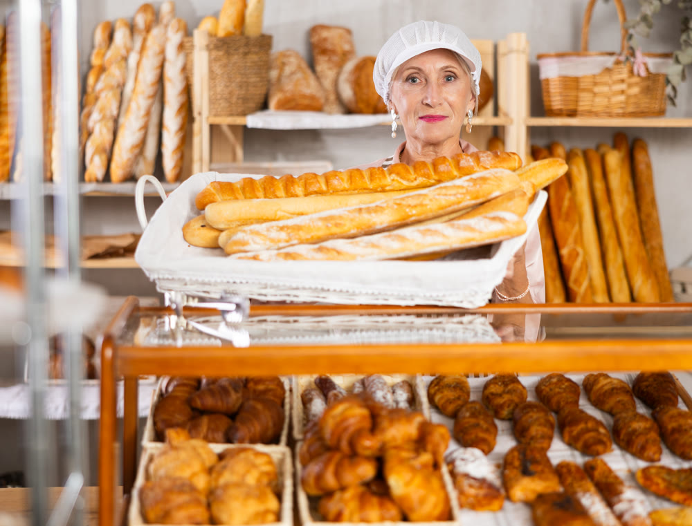Senior female baker worker sells baguette, shows many products