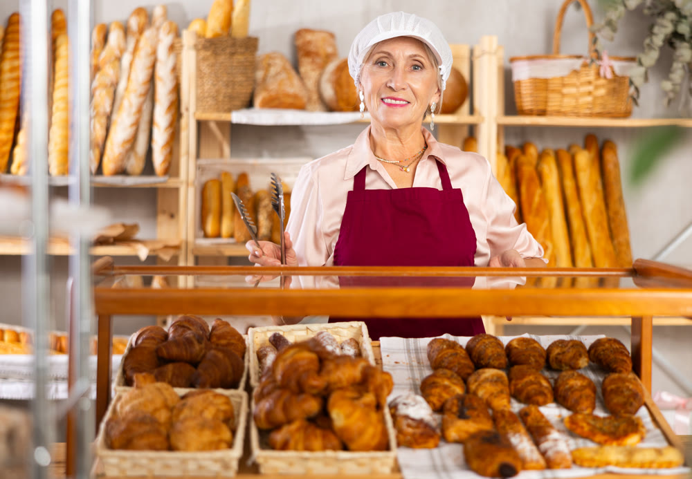 Portrait of elderly woman baker standing behind display case with fresh croissants. Offering to buy fresh baked goods