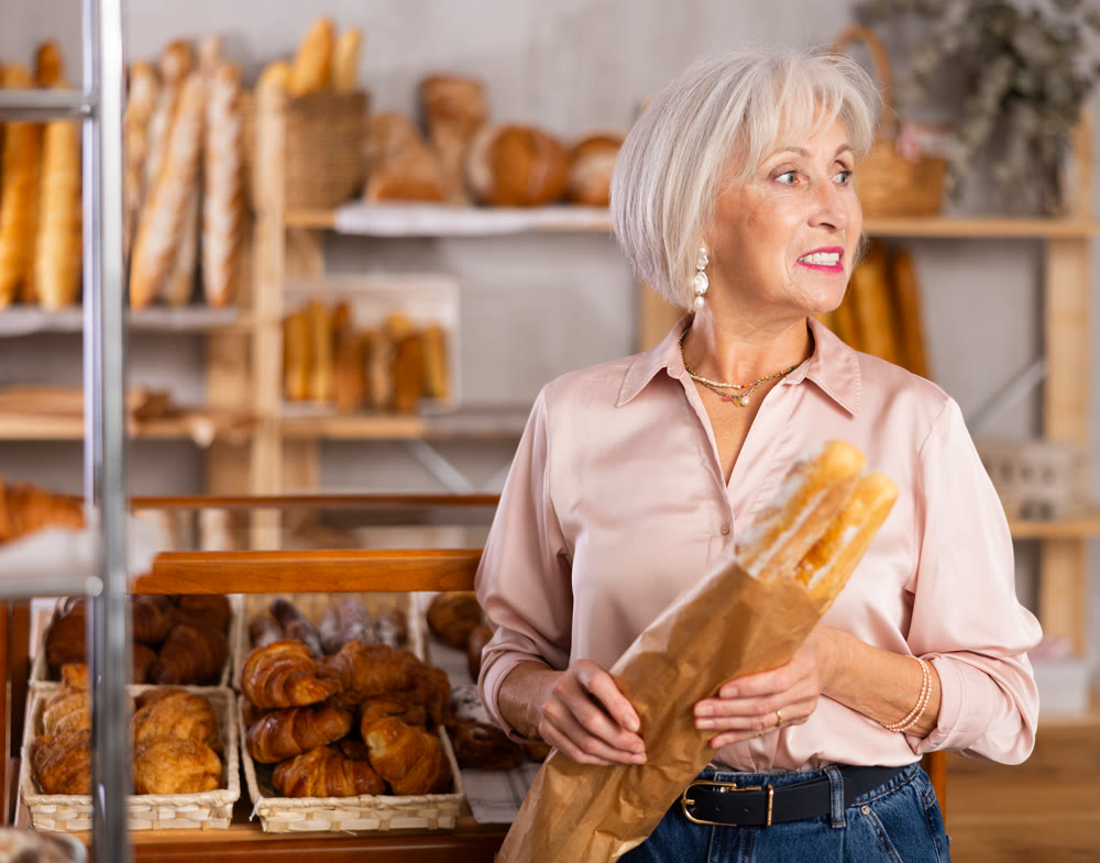 Elderly woman holds baguette wrapped in paper bag in bakery store