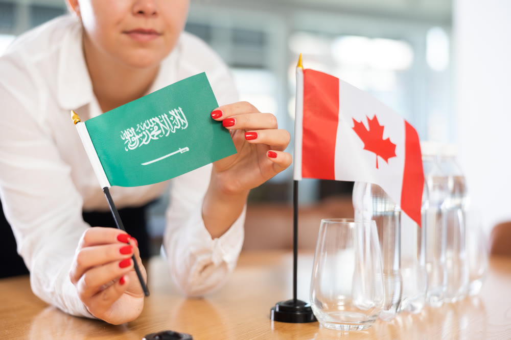 Young woman putting flags of Saudi Arabia and Canada on table in office