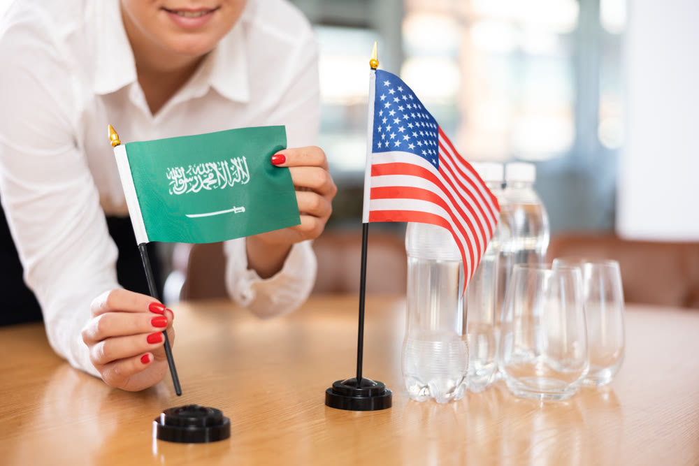 Young woman putting Saudi Arabia and USA flags on table in office