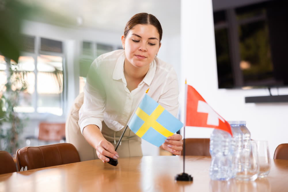 Young female assistant putting up swiss and swedish flags on table for international negotiations