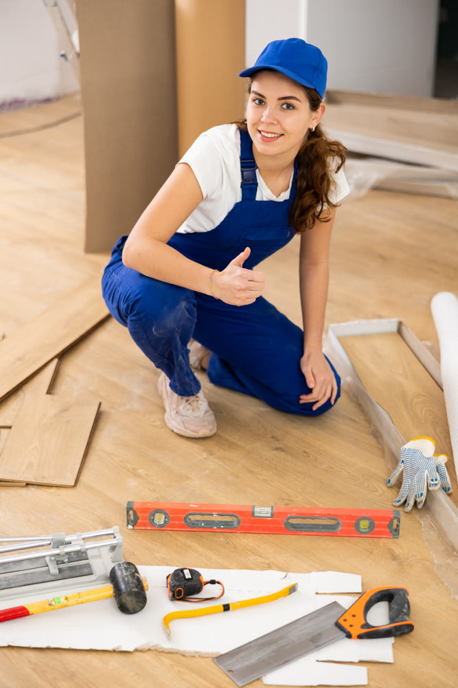 Young woman builder making thumb up gesture while installing parquet