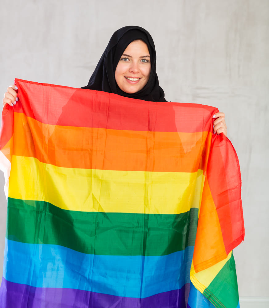 smiling Muslim woman in black hijab stands and holds striped pride flag in front of her.