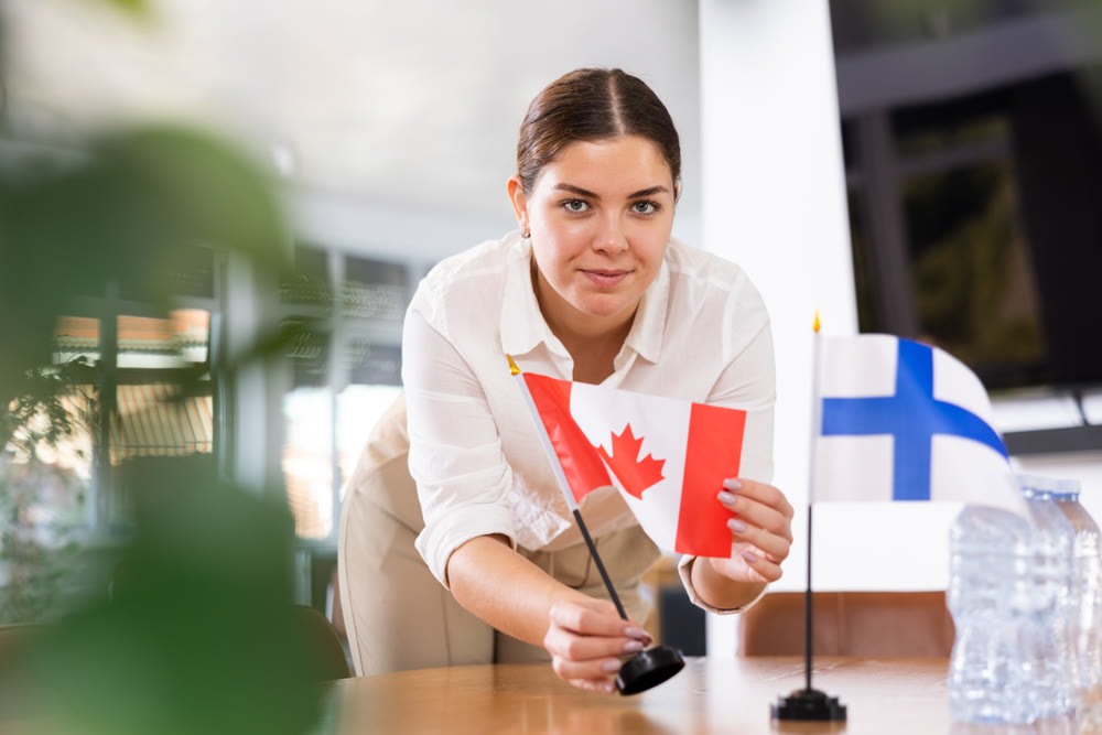 Positive young woman putting flag of Canada on the table with flag of Finland in conference room