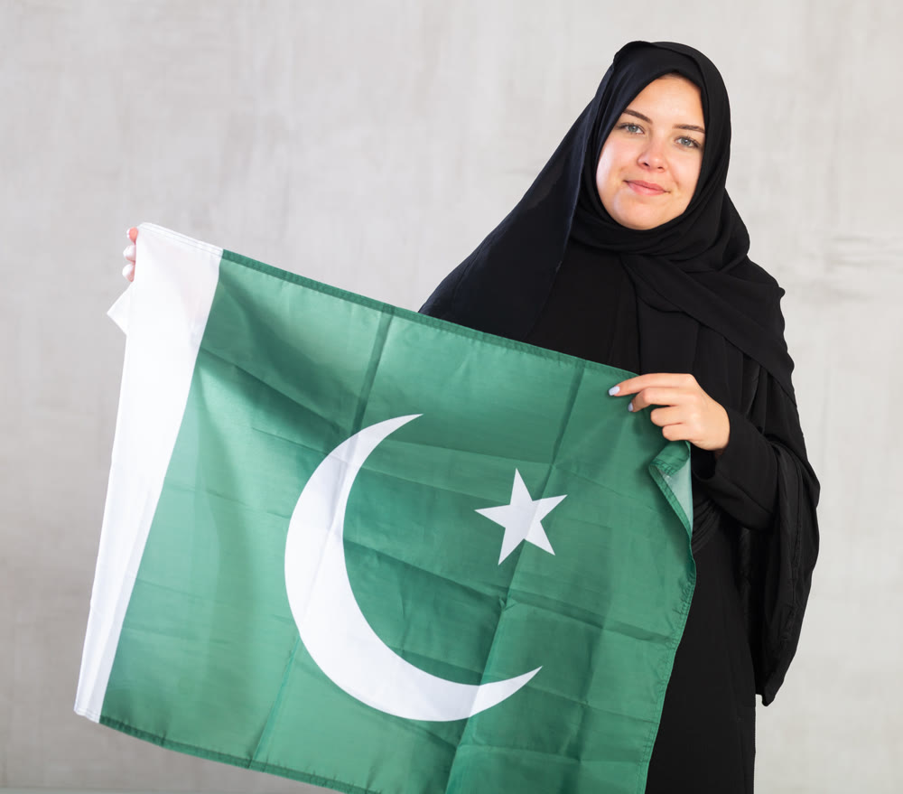 Balanced smiling Muslim woman in traditional black hijab holds flag of Pakistan