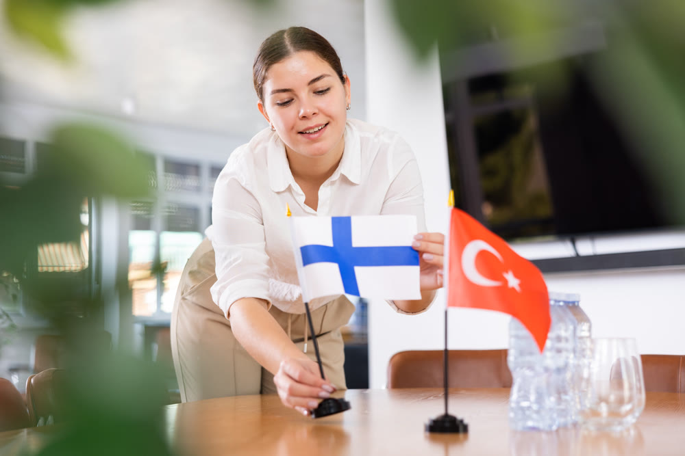 Positive young woman putting flag of Finland on the table with flag of Turkey in conference room