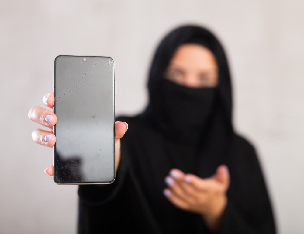 young Muslim girl with her face covered by burka shows empty dark smartphone screen.