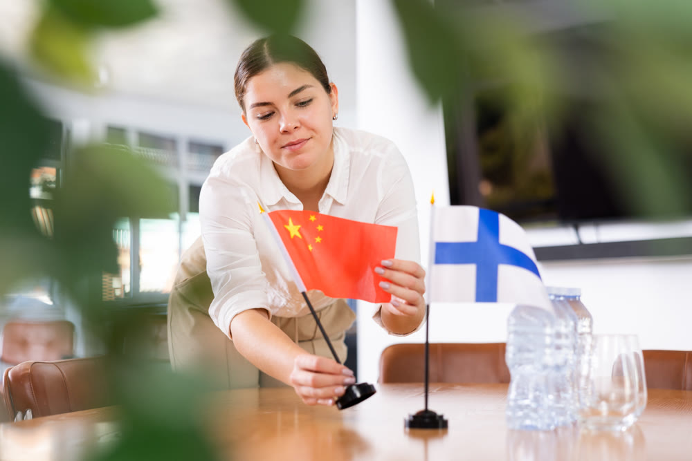Young female assistant putting up finnish and chinese flags on table for international negotiations