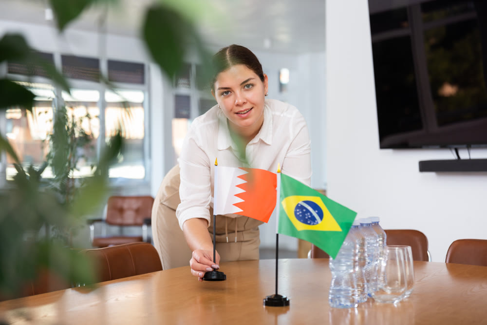 Positive young woman putting flag of Bahrain on the table with flag of Brazil in conference room