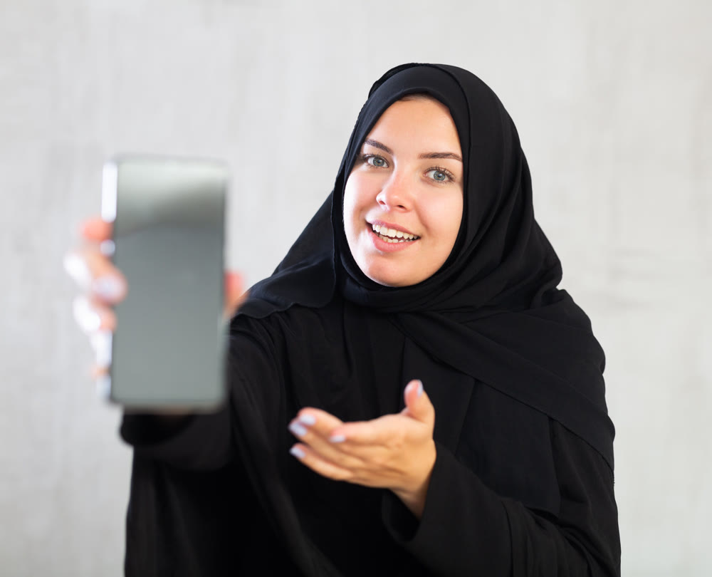 Smiling young woman dressed in traditional islamic clothes displaying brand new smartphone