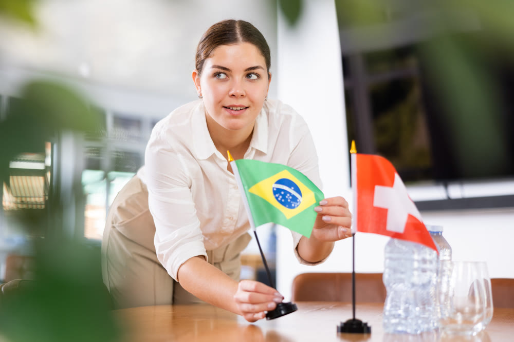 Young female assistant putting up swiss and brazilian flags on table for international negotiations