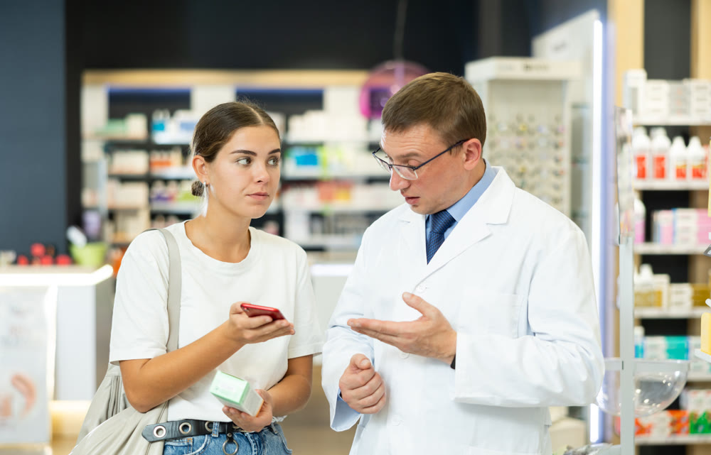 Positive male pharmacist helping young girl with electronic prescription in her phone choosing medical supplies at drugstore