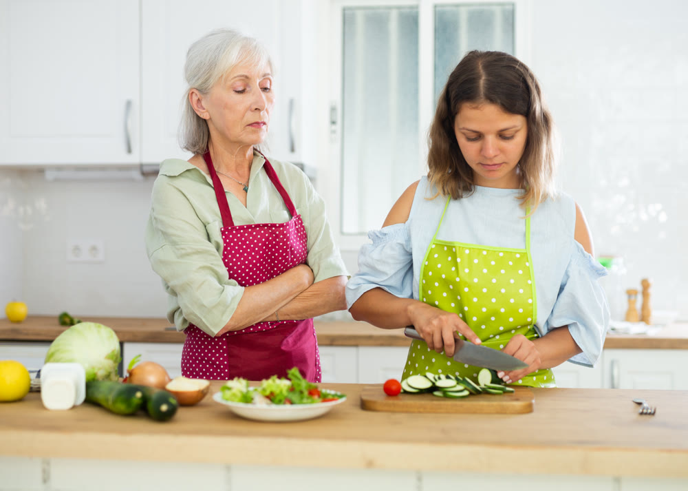 Old woman in apron observing her daughter cooking salad