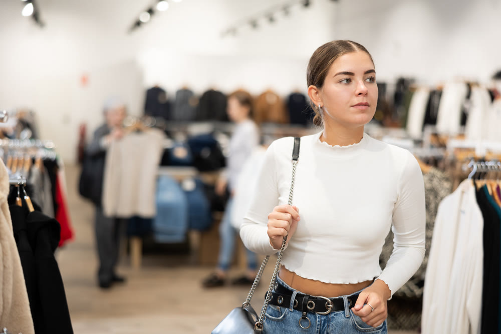 Positive joyful young woman looking for fashionable clothes in clothing boutique