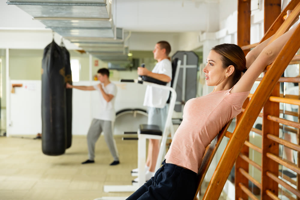 Young woman doing back stretching exercises on wall bars at gym