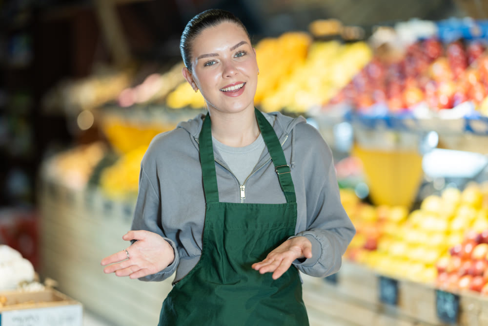 Young female shop assistant inviting in vegetable store