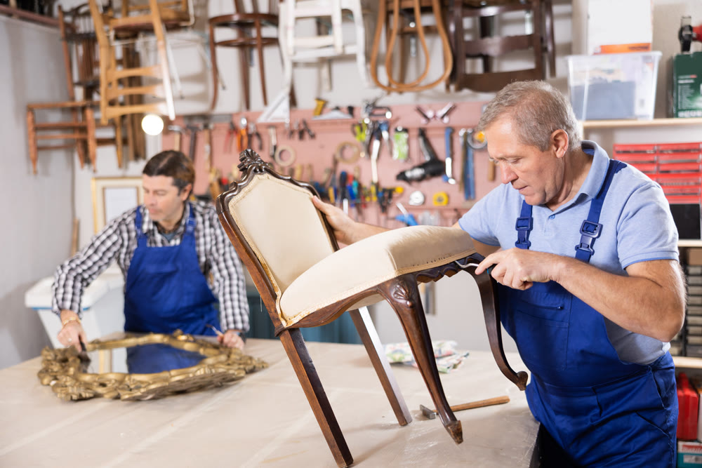 To replace upholstery, restorer removes old cloth seat covering from antique wooden chair.