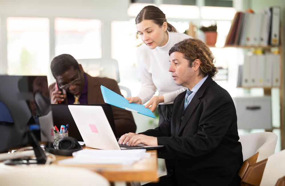 Woman with documents is standing next to male colleague with laptop