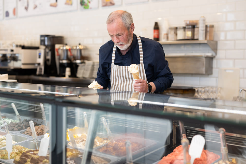 Elderly man in an apron offers ice cream at dessert counter with an inviting smile