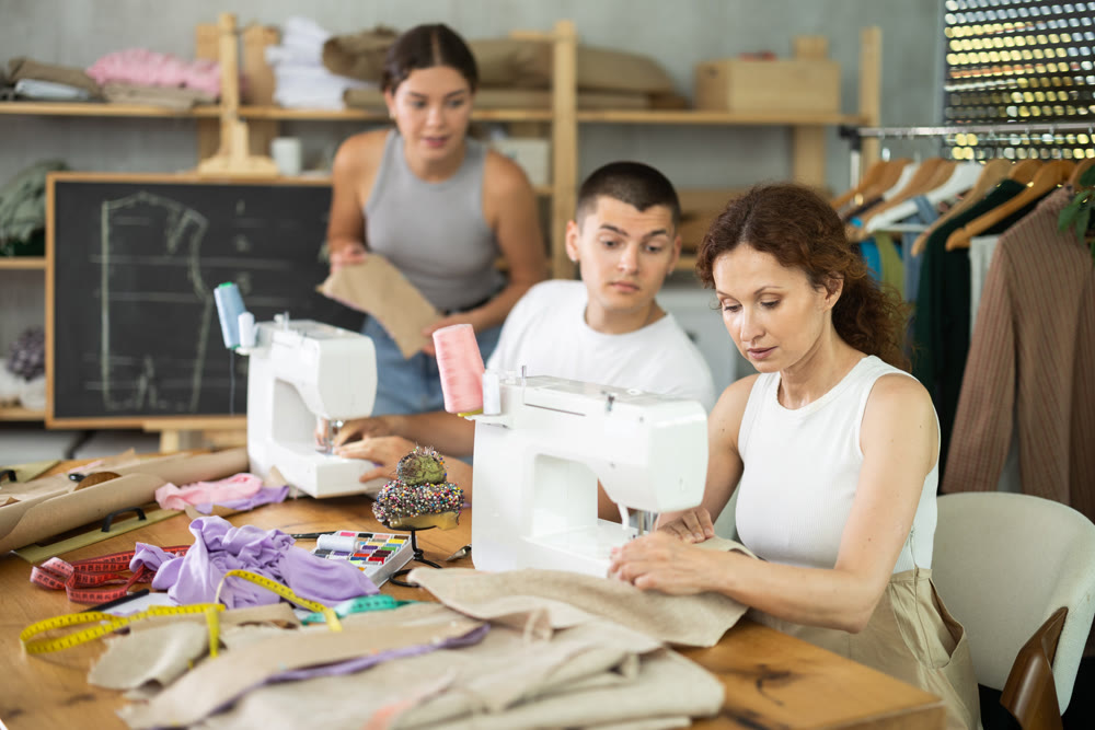 Multiethnic participants in coterie practice sewing, chatting, work textile and paper dress pattern