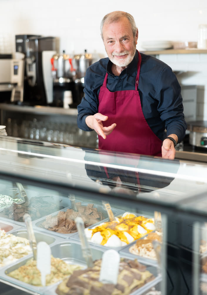 Elderly man in an apron offers ice cream at dessert counter with an inviting smile