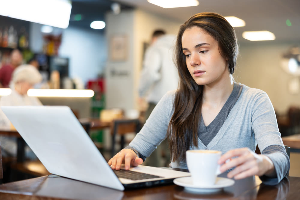 In cozy cafe hall, freelance young woman using laptop and drinks coffee.