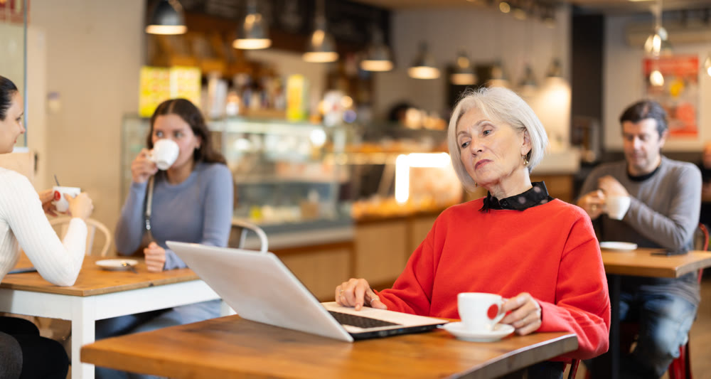 In cozy cafe hall, freelance senior woman works with laptop and drinks coffee.