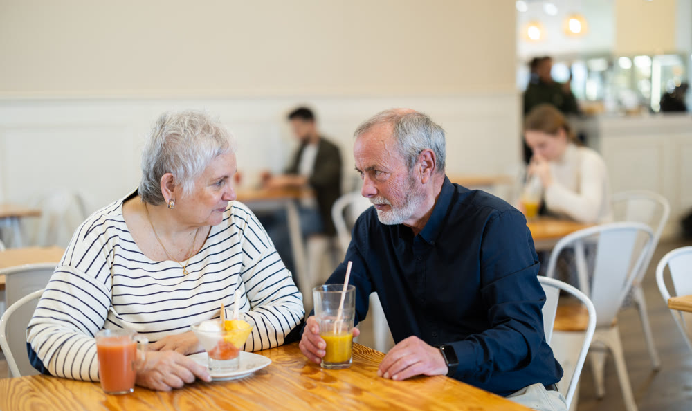 Elderly married couple are chatting happily with each other while sitting at table in a cafe and drinking juice and eat ice