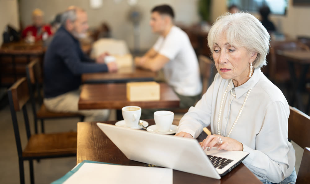 In cozy cafe hall, freelance senior woman using laptop and drinks coffee.