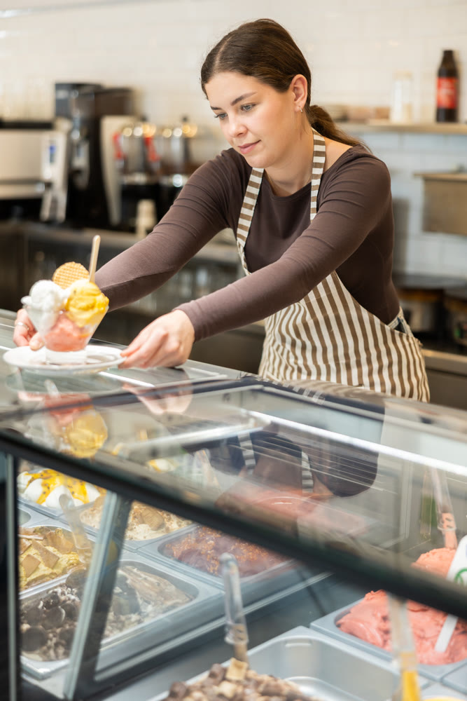 Pretty woman in an apron offers ice cream at dessert counter with an inviting smile