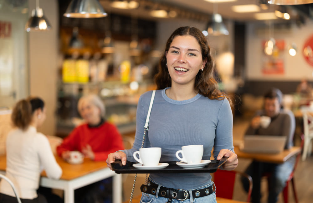 Girl visitor in self-service cafeteria carries tray with two cups of coffee.
