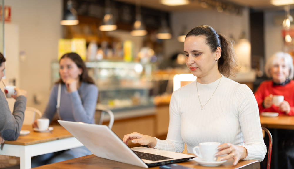 In cozy cafe hall, freelance woman works with laptop and drinks coffee.