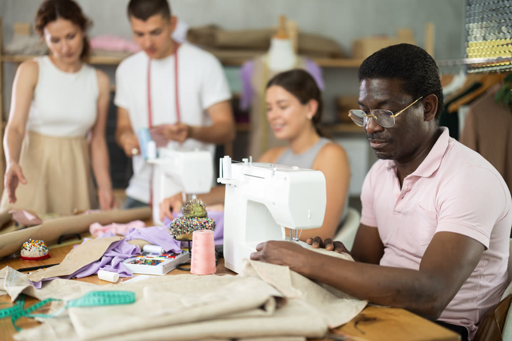Middle-aged man sewing while others working with sewing-machine and making details