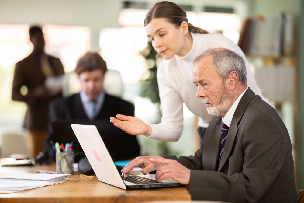 Experienced employee helping elderly worker with laptop