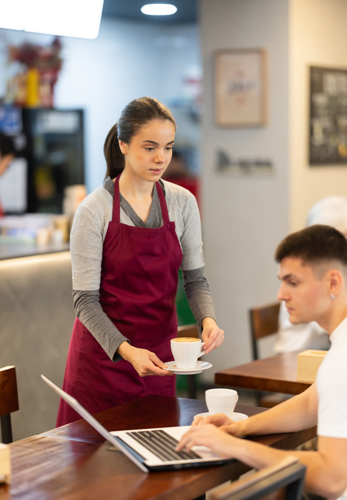 Young female waitress brings coffee to man with laptop