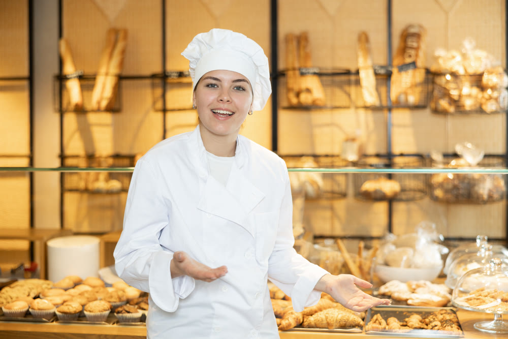 Female baker in a white coat and chef hat invites you to buy fresh bread and buns