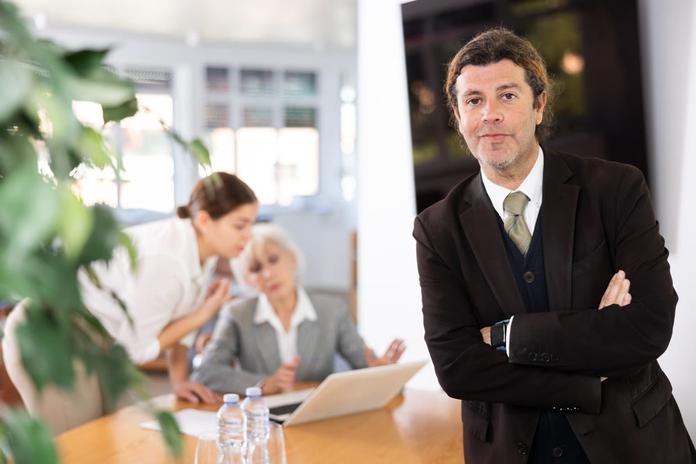 positive male colleague standing in meeting room and employees are talking behind him at table