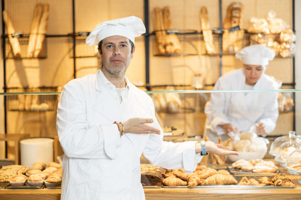 Successful baker in a white coat and chef hat invites you to buy fresh bread and buns